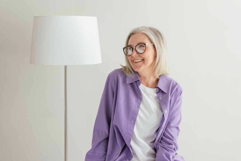 A joyful senior woman wearing eyeglasses and a purple shirt, sitting indoors next to a white lamp.