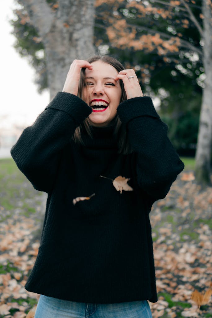 A woman in a black sweater joyfully laughing among fallen autumn leaves in a park.
