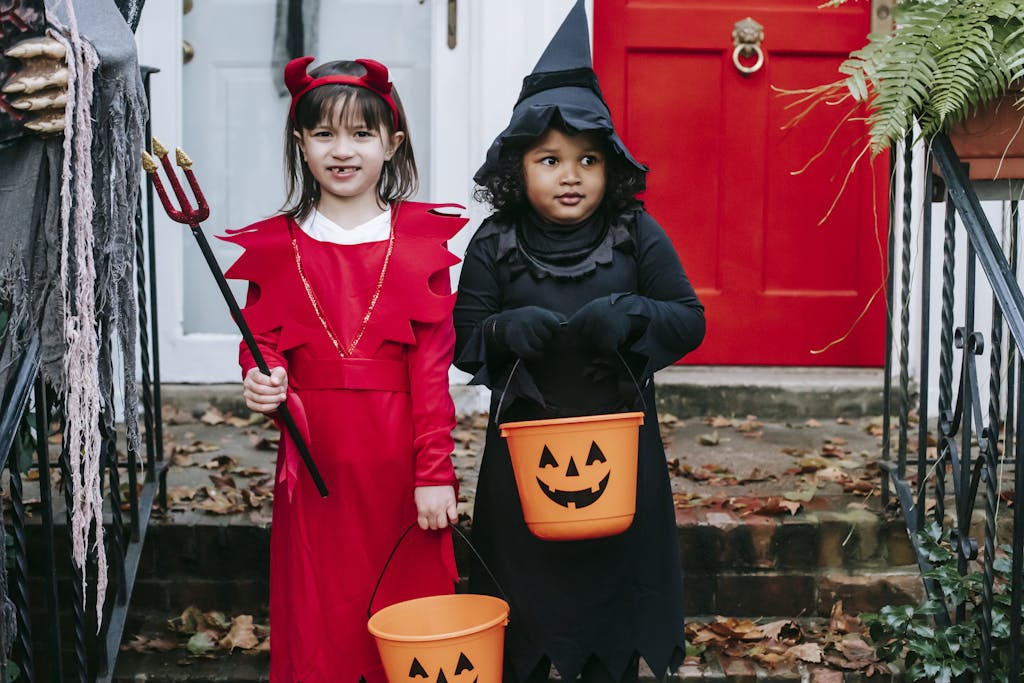Diverse preschool girlfriends in costumes holding buckets and trident while standing near door on Halloween